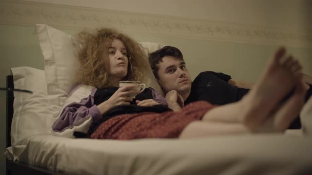 放松的夫妻躺在床上看电视，女人用热茶烫着舌头。无忧无虑的白人家庭周末在家里的卧室。婚姻休闲概念视频素材