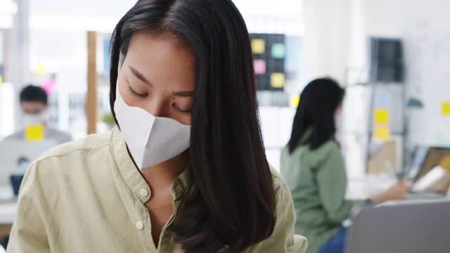 亚洲女企业家在新常态下戴着医用口罩进行社交距离以预防病毒，同时在办公室使用笔记本电脑工作。视频素材
