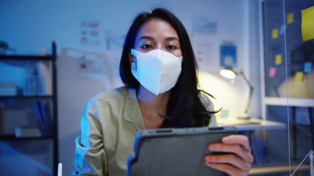亚洲商界女性在办公室工作时戴着口罩，在社交场合保持距离，以预防病毒，看着摄像机向朋友介绍计划的视频通话。视频素材