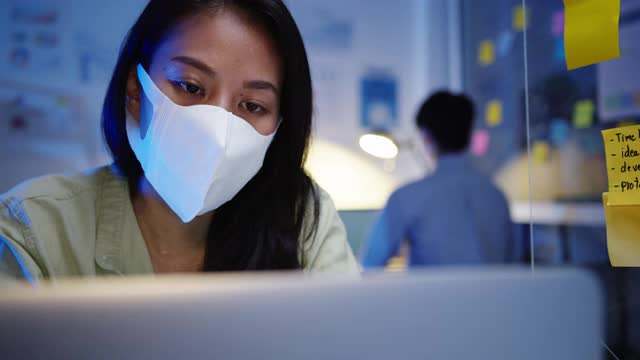 快乐亚洲女商人戴着医用口罩在新常态下进行社交距离以预防病毒，同时在晚上工作时使用笔记本电脑。视频素材