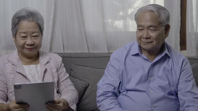 亚洲的祖父母和孙女在家里使用平板电脑。和年轻可爱的女孩一起躺在客厅的沙发上查看社交媒体，快乐地度过家庭放松时间。视频素材
