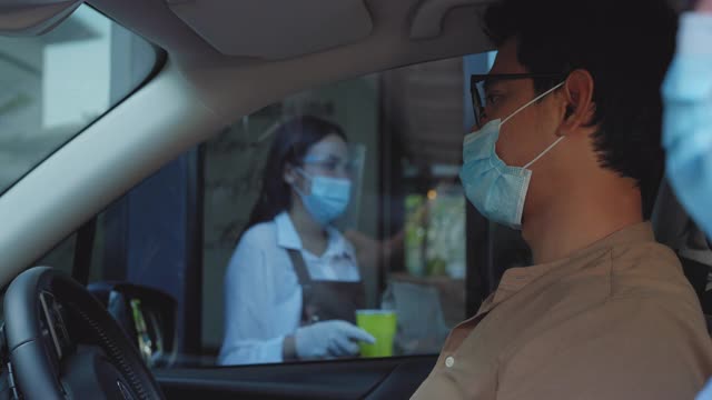 在冠状病毒爆发期间，亚洲男子戴着防护口罩，带着食品袋和咖啡，与女服务员在驾驶餐厅戴口罩和面罩。视频下载