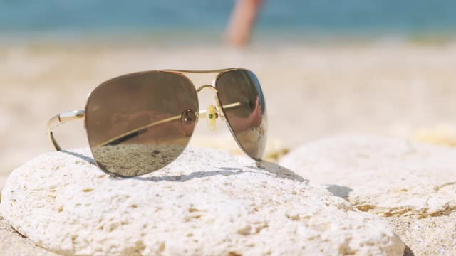 石头上的太阳镜反映出一个人躺在海滩上，建议海滩度假。视频下载