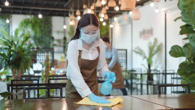 肖像有吸引力的亚洲餐厅员工戴口罩和面罩清洁桌子和地板用酒精和湿擦欢迎顾客。视频素材
