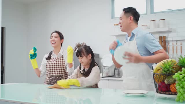 亚洲年轻家庭教他们的女儿清洁厨房柜台。孩子和父母一起跳舞，一起笑，很开心。可爱的家庭在快乐的打扫房间时的快乐时刻。视频素材