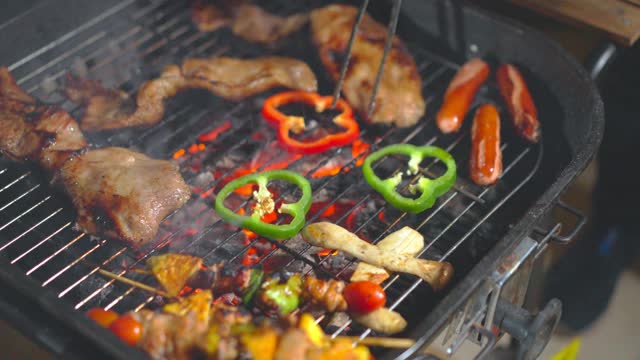 烧烤烤炉的特写镜头，有猪肉，甜椒，香肠和蘑菇在烤架上。人们用钳子把肉和蔬菜的另一面翻过来。户外活动与露营概念。视频素材