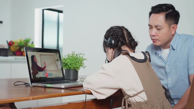 由于冠状病毒大流行，亚洲小女孩通过数字远程互联网会议从学校老师那里学习在线课程。孩子看着电脑笔记本电脑屏幕坐在父亲。视频素材