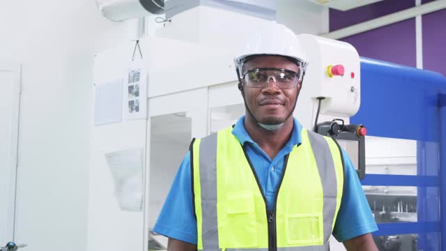 非裔美国男工程师的肖像，微笑着站在生产工厂看着相机。男工程师在生产过程中通过监督员工操作机器来照顾健康的工作。视频素材