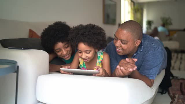父亲和女儿一起躺在家里的沙发上使用电子平板电脑视频素材