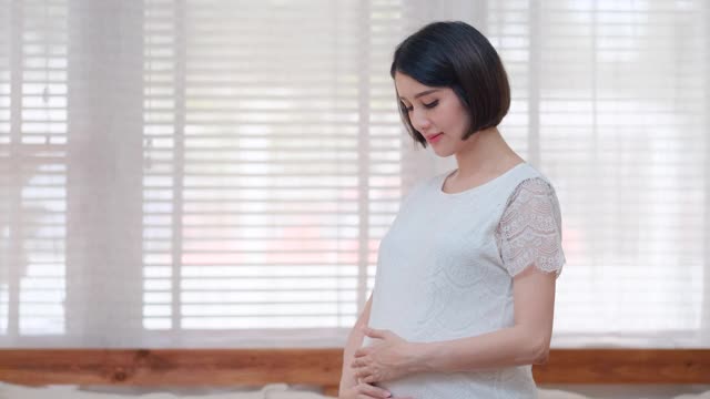 亚洲孕妇站在客厅，环顾四周，触摸她的肚子，以表达对她的孩子的爱和关心的情感视频素材
