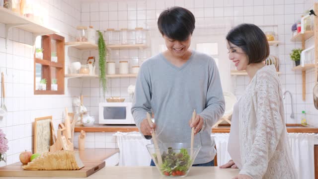 亚洲男人或丈夫准备沙拉在玻璃碗站和他怀孕的妻子，他们看起来很高兴在厨房烹饪视频素材