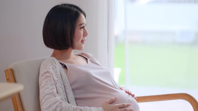 侧面的亚洲孕妇坐在椅子前玻璃门，她透过白色窗帘，摸她的肚子视频素材