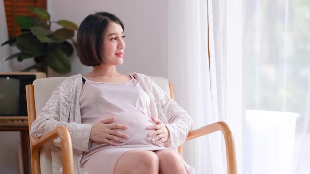 亚洲美丽孕妇坐在椅子上，在玻璃门前摸着肚子，用温暖的晨光环顾四周，表达幸福视频素材