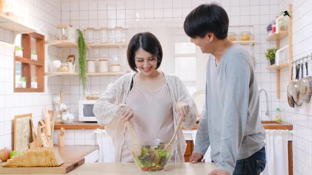 亚洲怀孕的妻子女人准备沙拉在玻璃碗和她的丈夫，他们看起来很高兴在他们家的厨房烹饪视频素材