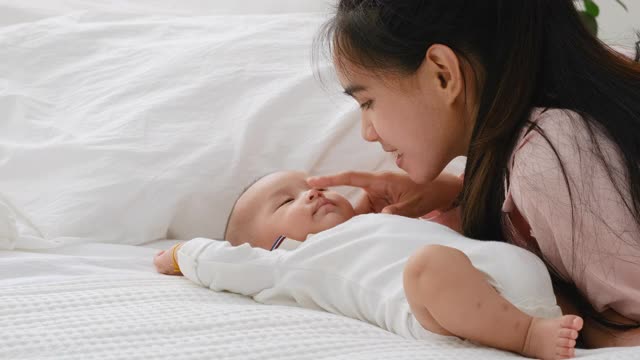 年轻的亚洲母亲逗弄躺在床上看起来睡着的新生儿，用她的手指触摸婴儿的鼻子与晨光视频素材
