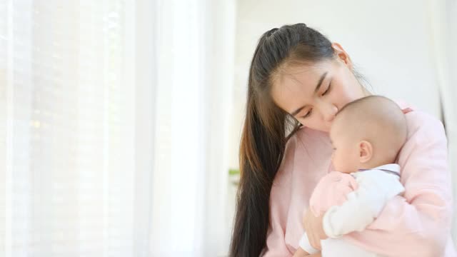 亚洲母亲抱着她的新生儿在她的胸部，环顾四周的白色窗帘，享受呆在家里视频素材