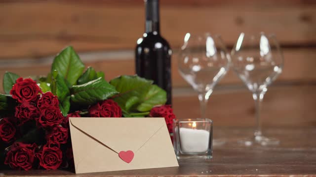 桌上有一瓶酒和两个杯子，旁边点着一支蜡烛。一束红玫瑰，一个女人的手拿着一个心形的信封。情人节节日的概念,视频下载
