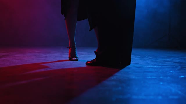 一个男人和一个女人的脚的剪影在一个黑暗的房间和明亮的蓝色和红色聚光灯跳舞。双脚在黑暗的地板上跳着拉丁舞。视频素材
