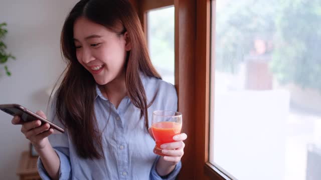 可爱的少女或妇女的肖像使用手机和喝橙汁在玻璃窗户前，看起来在家里很快乐视频素材