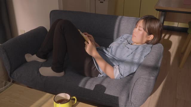 年轻女子喝热饮，坐在沙发上放松，在家里的客厅看书视频素材