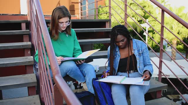 两名年轻的多民族妇女正坐在大学校园的木楼梯上学习视频下载