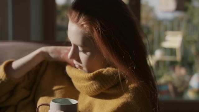 年轻女子在阳光下在家的沙发上喝茶视频下载