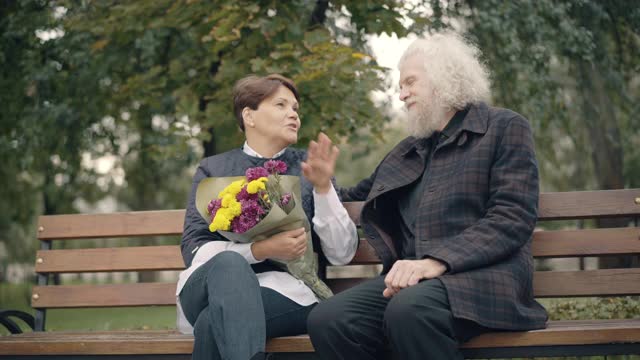 积极放松的一对白人退休人员坐在长椅上的秋天公园聊天。无忧无虑的快乐男女，享受户外的快乐休闲。爱情和约会的概念。视频素材
