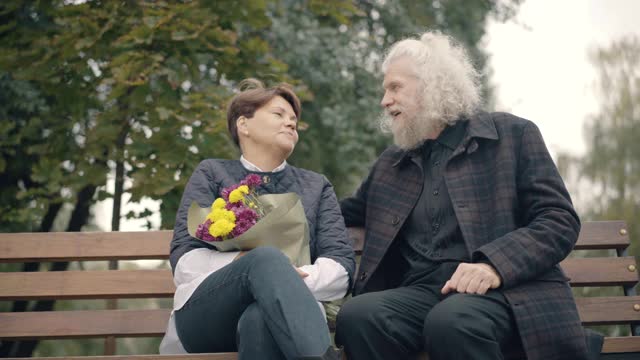 正面英俊的老男人与女人坐在长椅上聊天在秋天公园。爱的白人丈夫与妻子在户外捧着一束花约会的肖像。爱和休闲。视频素材