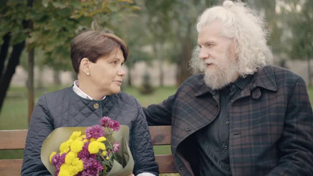 一对轻松的老夫妇坐在长椅上，在户外享受着春日或秋日。正面的白人退休男女聊天和微笑的肖像。约会和爱情的概念。视频素材
