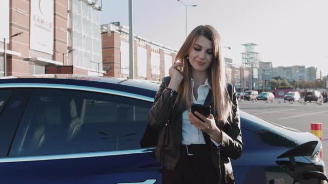 欧洲女性在等待时使用智能手机，电源连接到电动汽车上为电池充电。电动汽车充电视频素材