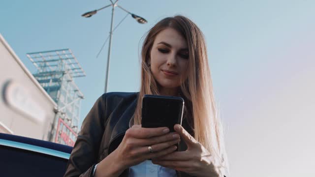 兴奋的年轻女子拿着手机，在约会app上聊天感到开心站在街道附近的车，女孩享受手机在线投注游戏胜利成功在手机上读到喜讯视频素材
