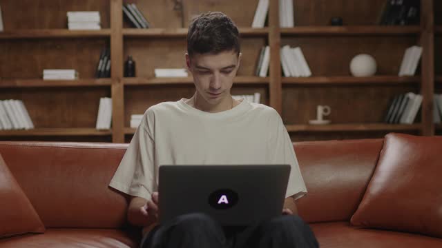 20多岁的年轻人坐在沙发上使用笔记本电脑。男性自由职业者在笔记本电脑上打字，上网。千禧一代在家学习。在线工作和教育。视频素材
