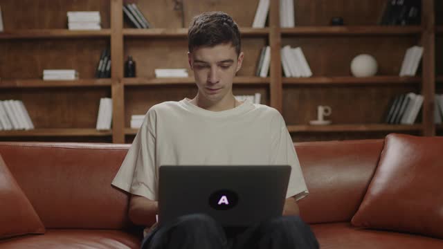 20多岁的年轻人坐在沙发上用笔记本电脑打字。男性自由职业者关闭笔记本电脑和思考。千禧一代在家学习。在线工作和教育。视频素材