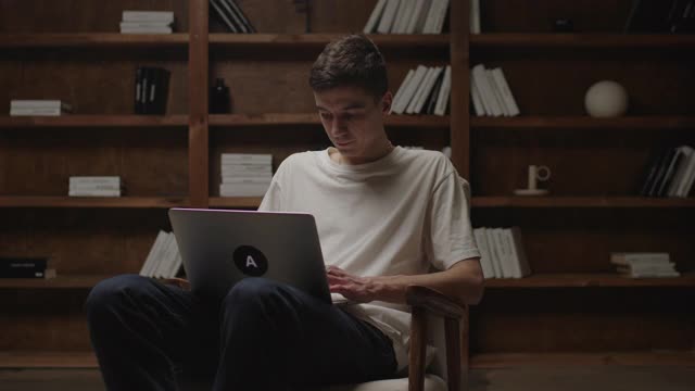 20多岁的年轻人坐在扶手椅上在笔记本电脑上打字。千禧一代在家图书馆学习。在线工作和教育理念。视频素材