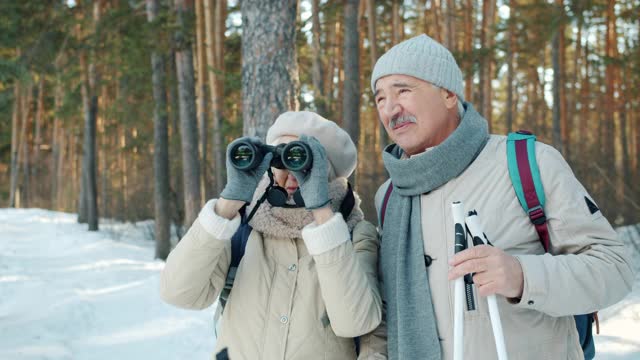 在冬季公园里，老妇人拿着滑雪杖，通过望远镜和丈夫交谈，观察风景视频下载
