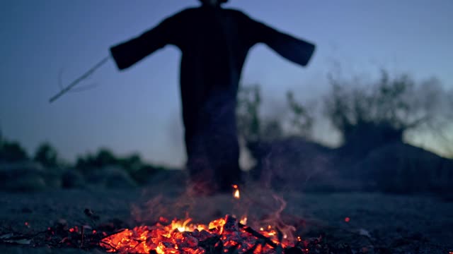 穿着黑色长袍的幽灵死在火旁。地上有余烬，神秘人物用木棍做萨满教。万圣节的一天。视频素材