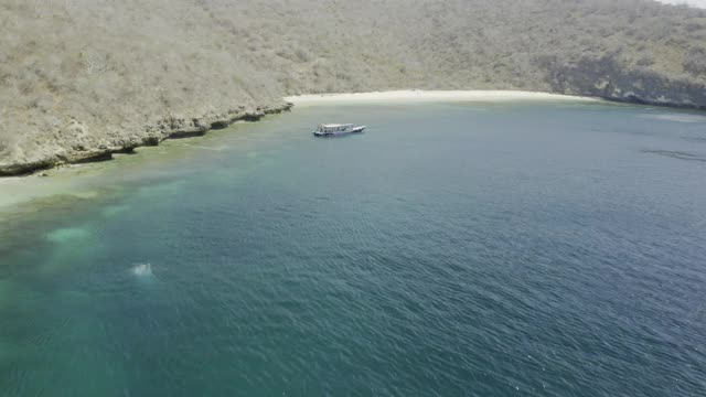 印尼库塔，一架引人注目的航拍向前，一艘船停泊在绿松石色的海湾上，周围环绕着杂草丛生的山丘和沙滩视频下载