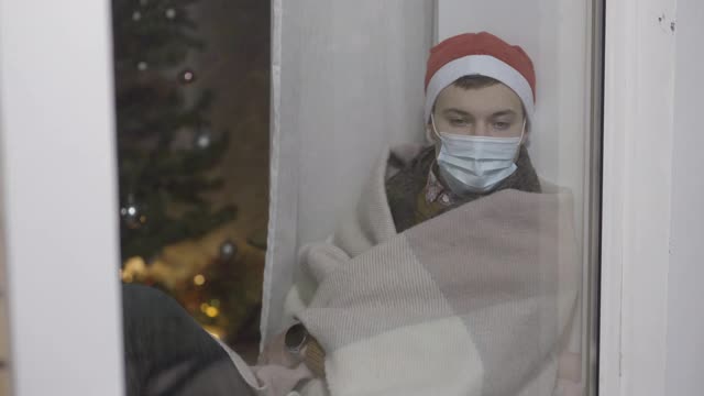 一名疲惫的年轻人戴着口罩和圣诞帽，坐在窗台玻璃后面睡着。一个疲惫不堪的白人在家里过新年的肖像。孤立和封锁视频下载