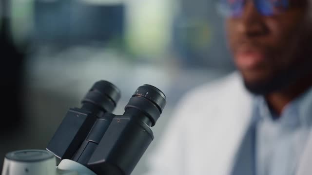 一个英俊的黑人男性科学家戴眼镜和在显微镜下看的宏近镜头。在现代实验室用技术设备研究分子样本的微生物学家。视频素材