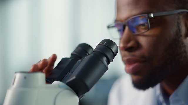 一个英俊的黑人男性科学家戴眼镜和在显微镜下看的宏近镜头。在现代实验室用技术设备研究分子样本的微生物学家。视频素材