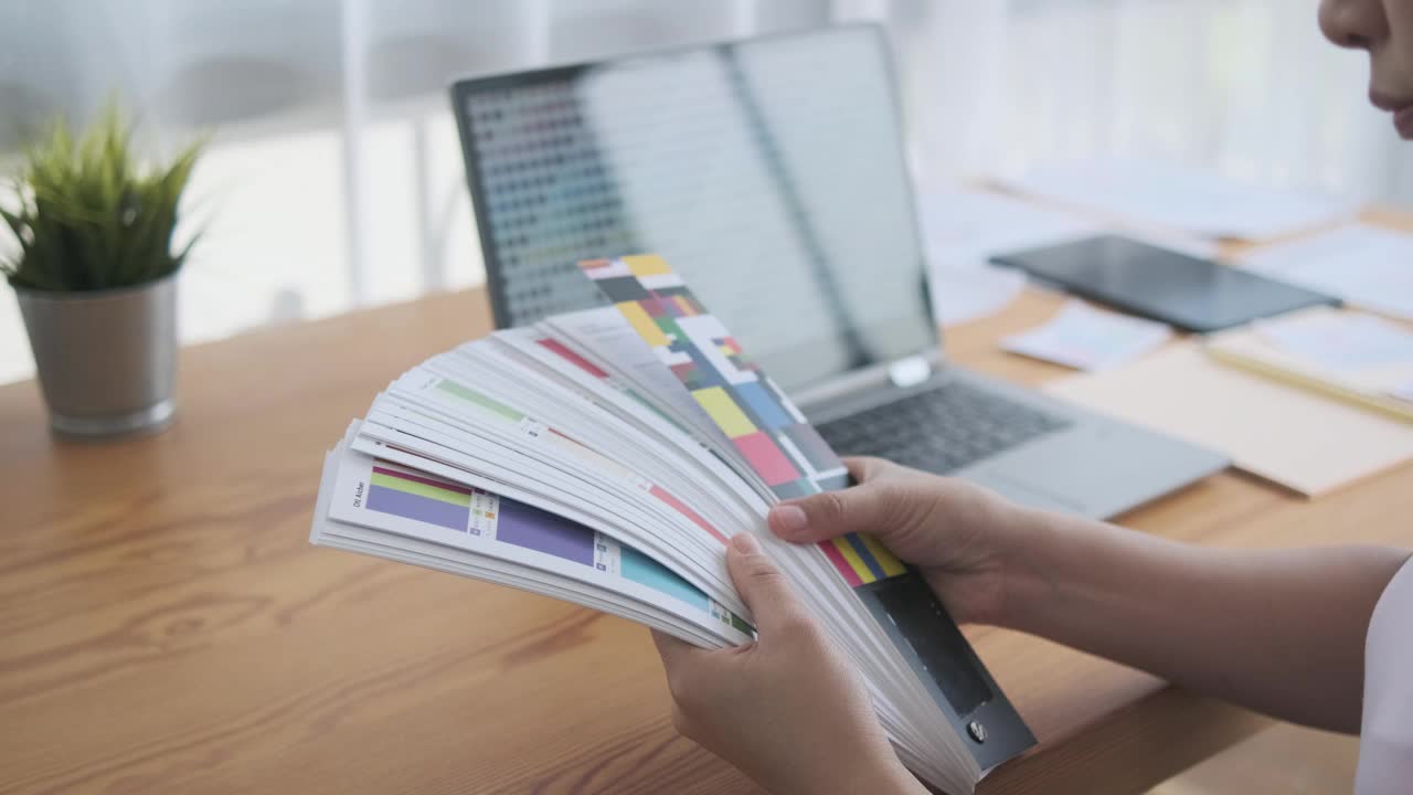 亚洲女性设计师选择颜色样本托盘从图表条创造性的装饰工作与色彩和谐，在办公室工作空间与电脑笔记本规划项目室内设计。视频下载