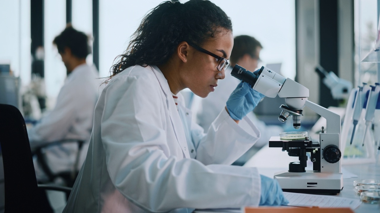 医学实验室:美丽的黑人科学家在显微镜下观察，写下分析数据。年轻的生物技术专家，使用先进的设备。侧视图慢动作放大视频素材