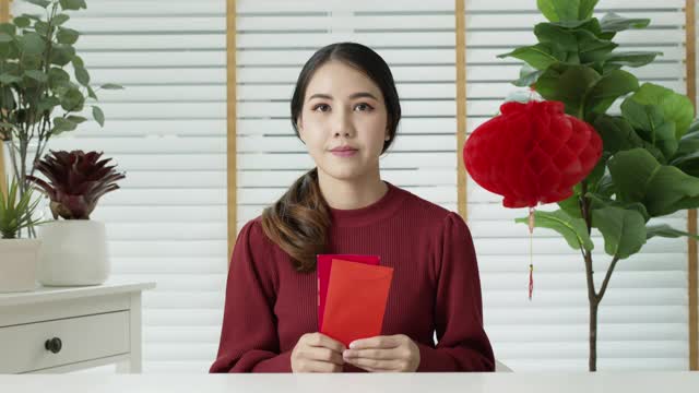 自拍视频通话中，一位迷人的年轻亚洲女士穿着红色的中国新年庆祝服，开心地微笑着交谈，看着拿着红包的手机。视频素材