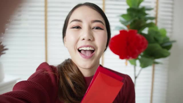 自拍视频通话中，一位迷人的年轻亚洲女士穿着红色的中国新年庆祝服，开心地微笑着交谈，看着拿着红包的手机。视频素材