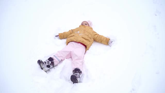 一个蹒跚学步的小女孩在冬天穿着冬衣做雪天使。和孩子们一起进行户外活动。真实的生活。快乐的童年视频下载