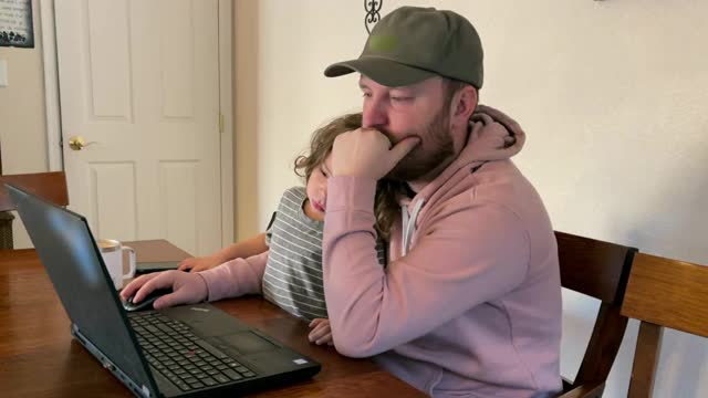 一位年轻的父亲在家里用笔记本电脑工作，他三岁的儿子坐在他的腿上看他工作视频下载