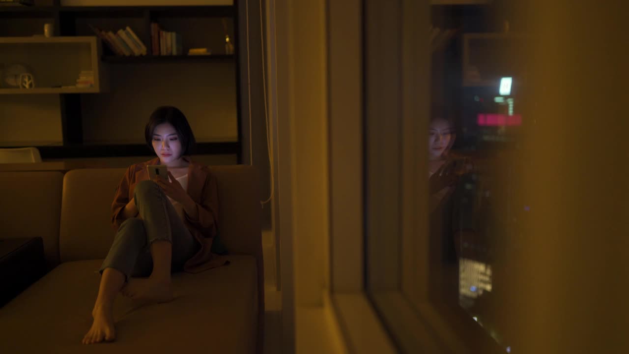 年轻的亚洲华人女子在晚上的客厅里用手机上网视频素材