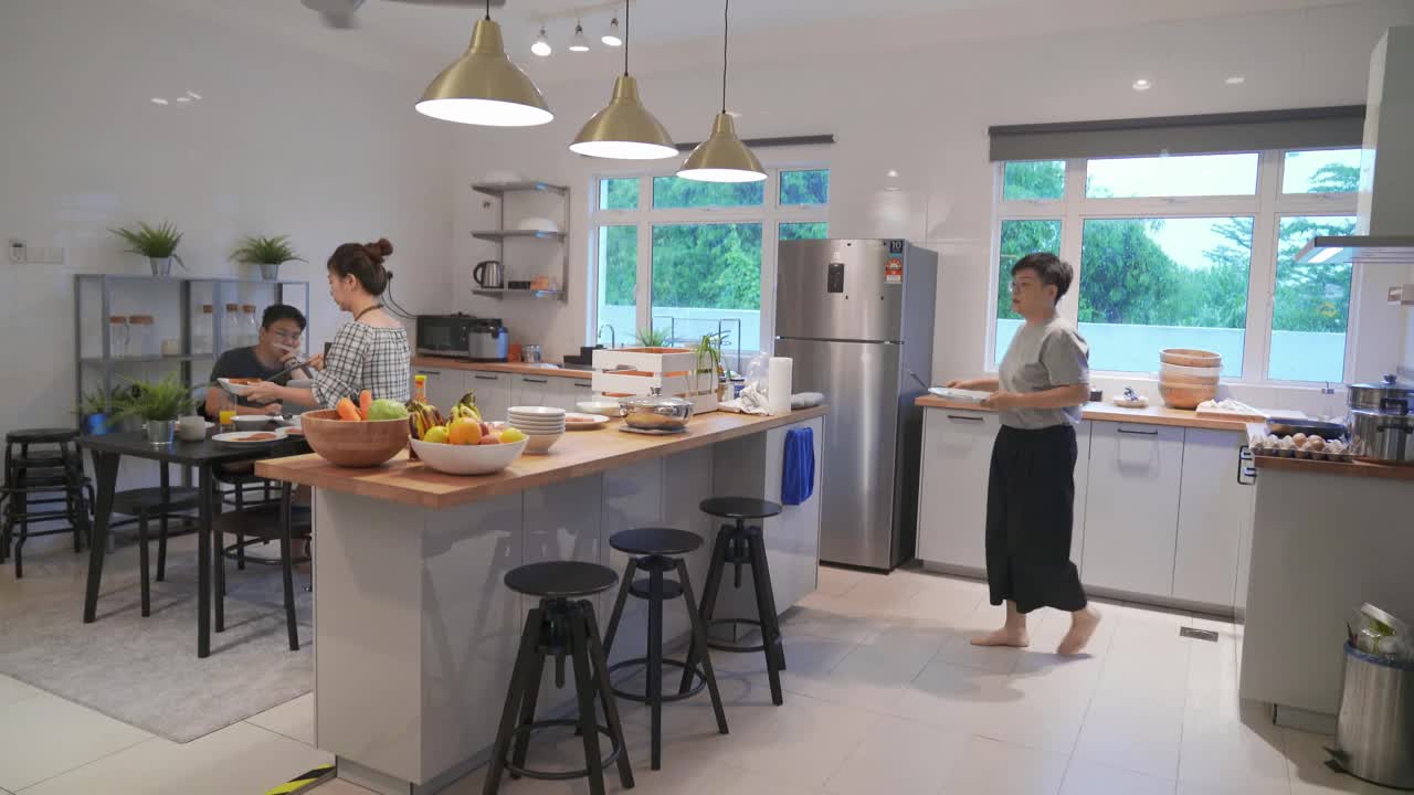 亚洲华人男子在家里用笔记本电脑工作，而家庭主妇和姐姐在厨房准备早餐视频下载