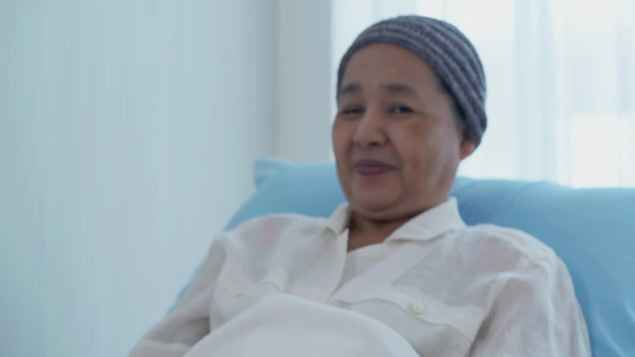 62岁的亚洲老年妇女微笑着，头部覆盖着一块因化疗导致的癌症的布，看着医院里的摄像头，她拿着绿色背景的手机。绿屏新技术概念。视频素材