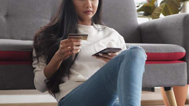 年轻的亚洲女性用智能手机和信用卡支付网上购物，坐在家里的沙发上。数字时代的生活方式视频素材